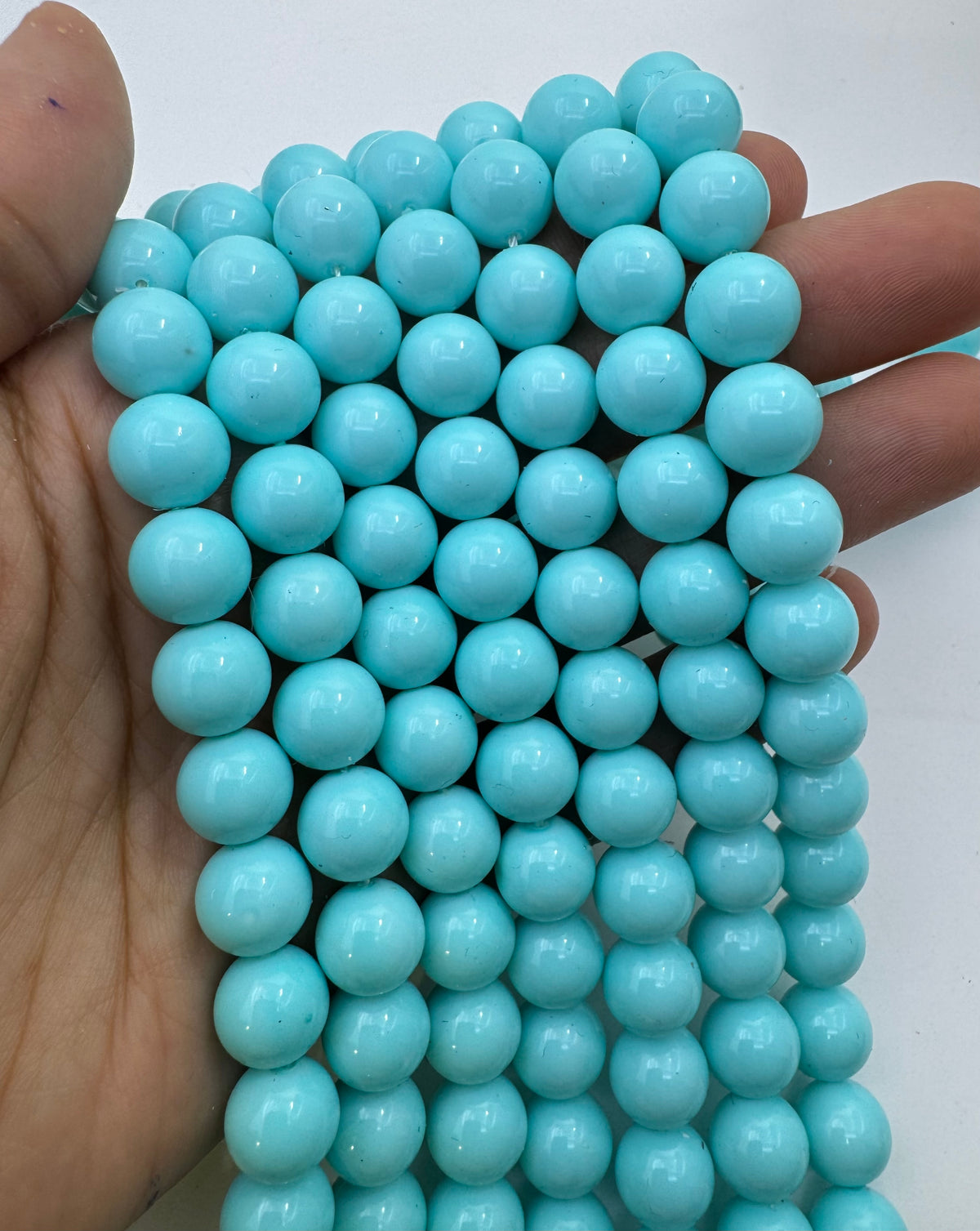 A hand holding a bundle of light blue turquoise shell pearl round beads against a plain white background.