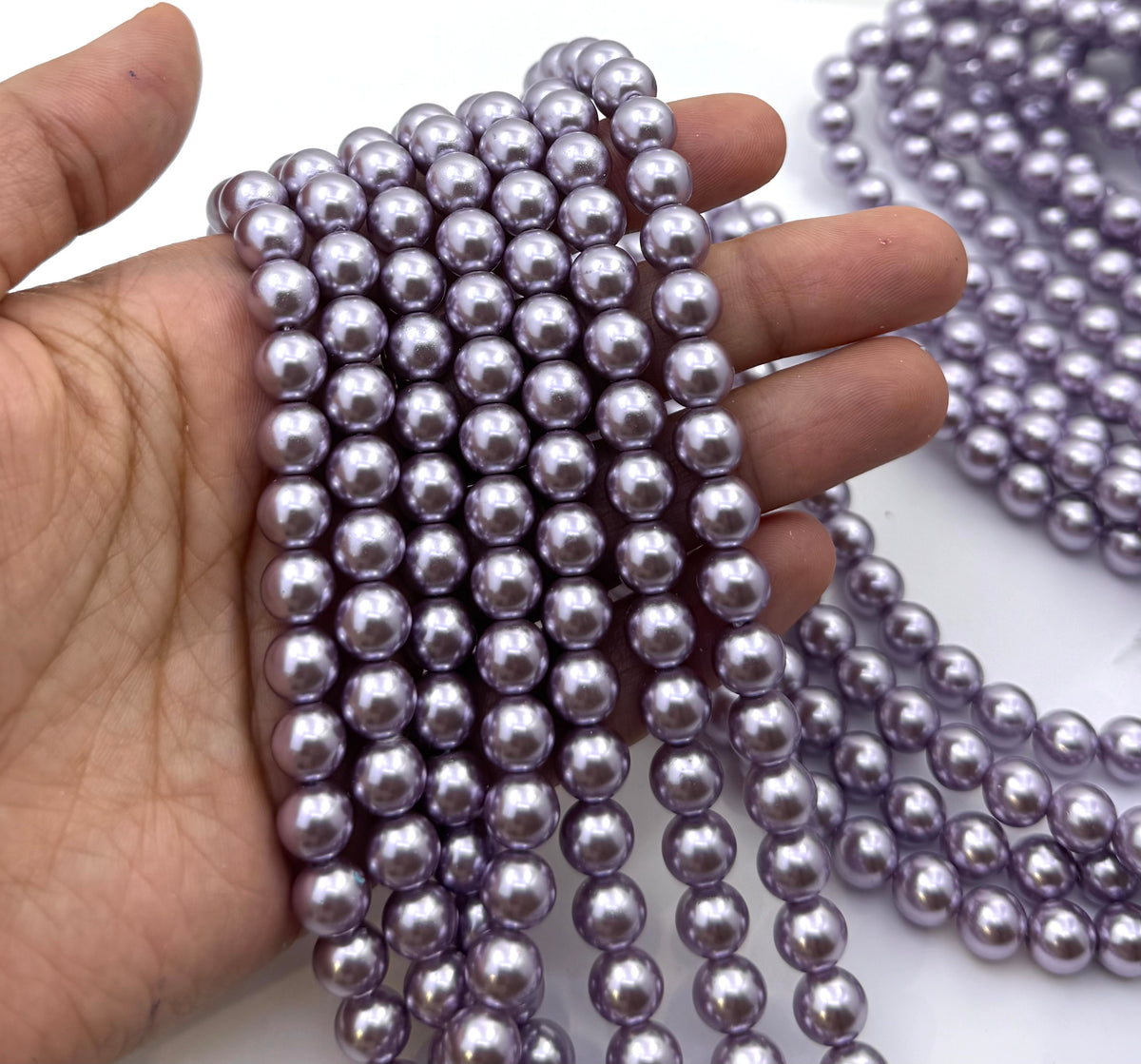 A hand holding multiple strands of glossy, lavender-purple shell pearl beads, with additional strands visible in the background on a white surface.