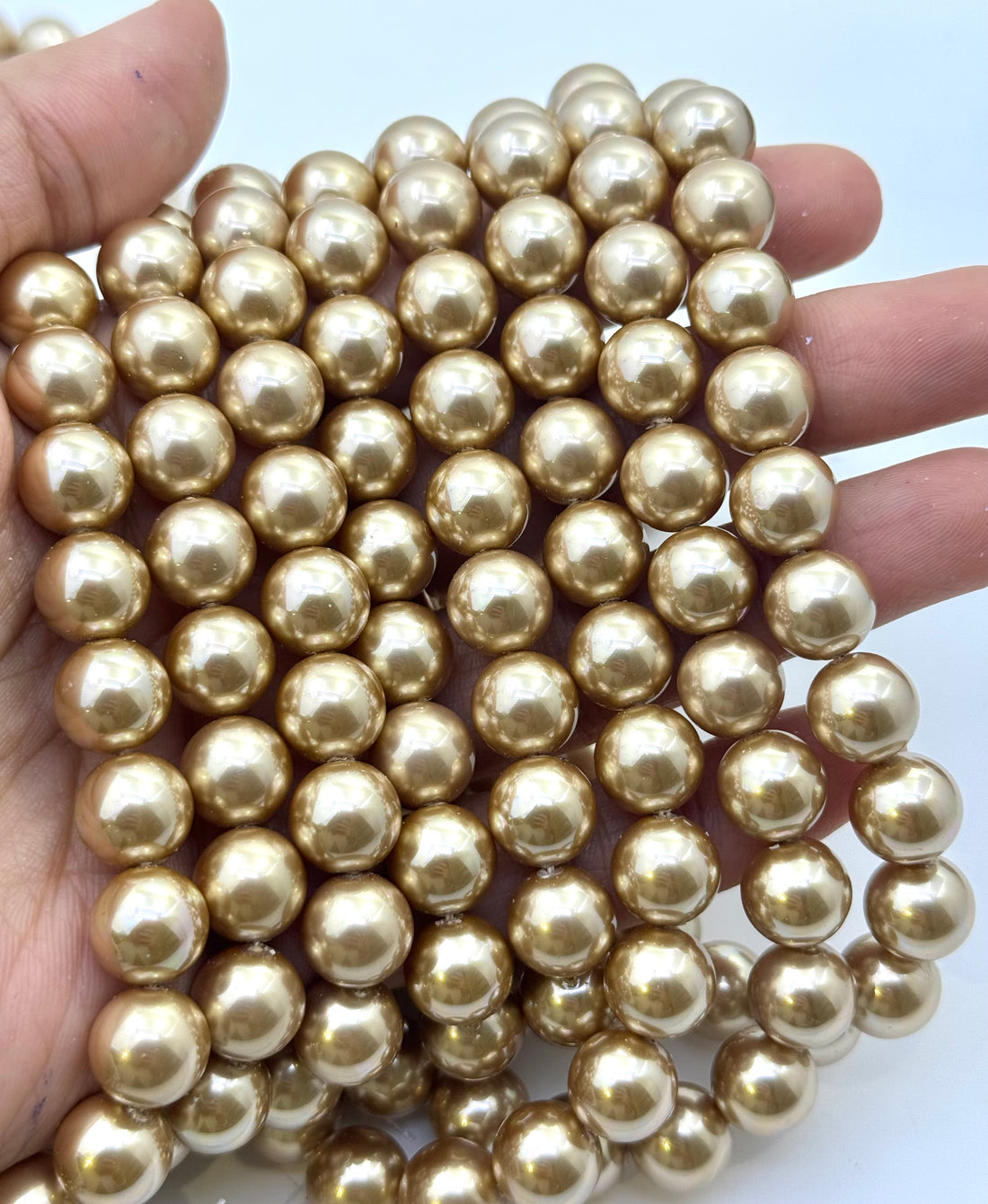 A hand holding a dense bundle of shiny metallic gold shell pearl round beads against a plain light background.