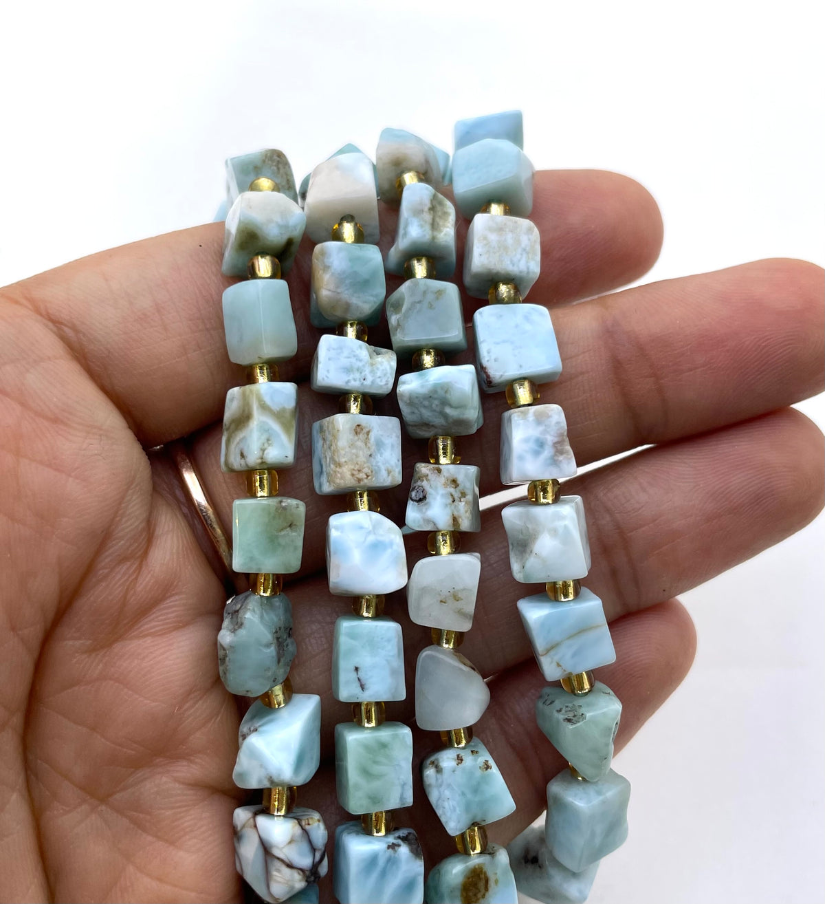 A hand holding three strands of raw, irregular larimar beads separated by small gold spacer rings, set against a plain white background.
