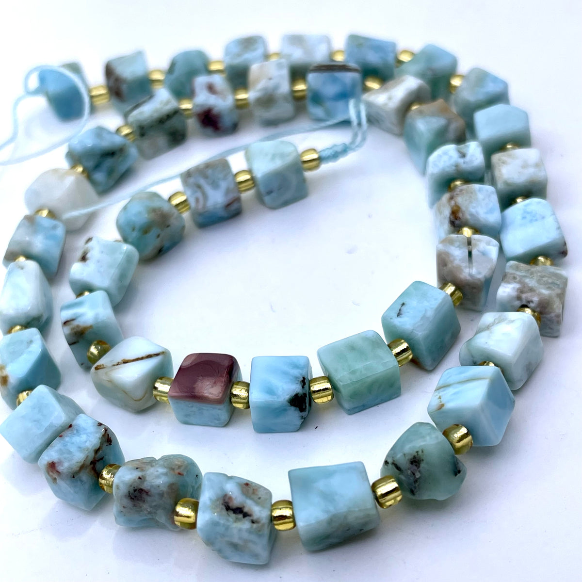 Light blue larimar gemstone cube beads with gold spacer rings, arranged in a spiral on a white background.