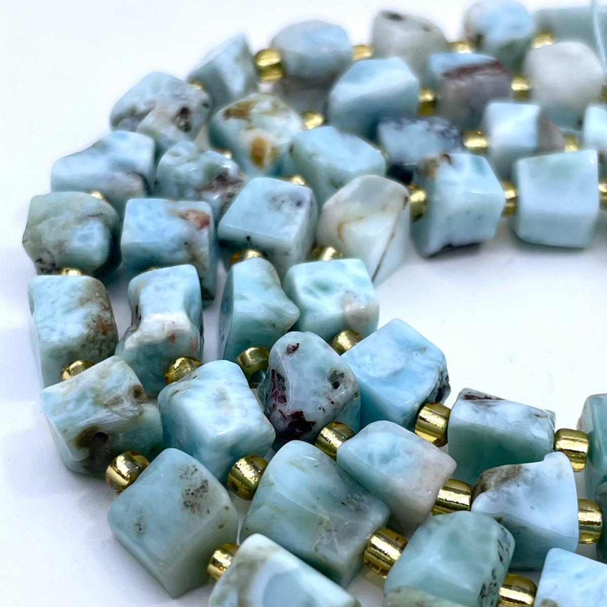 A close-up of a strand of raw, irregularly shaped light blue larimar beads separated by small gold spacer beads, arranged on a plain white surface.