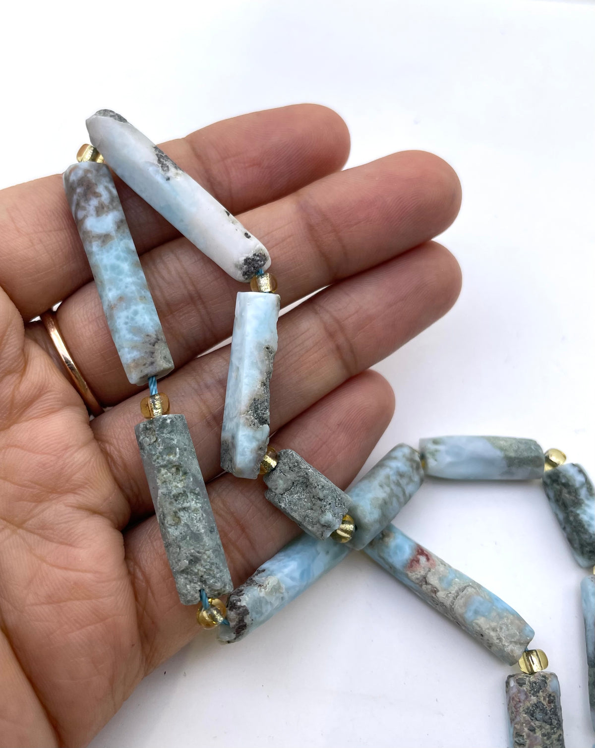 A hand holding a strand of natural larimar marble cylindrical beads with gold spacer rings, shown against a plain white background.