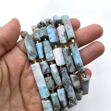 A close-up of a hand holding tube beads made of natural baroque-style larimar in light blue, white, and gray, separated by small gold spacer beads, against a plain white background.