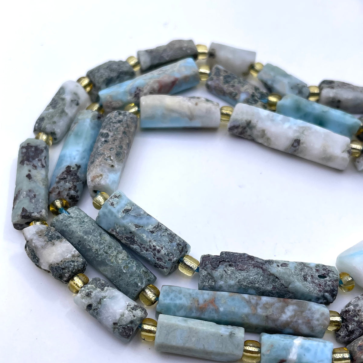 A strand of cylindrical larimar stone beads in natural blue, gray, white, and brown marbled patterns, separated by small gold spacer rings, photographed on a white background.