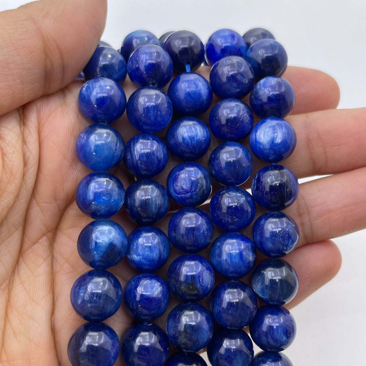 A hand holding a strand of polished round kyanite blue beads, against a plain white background.